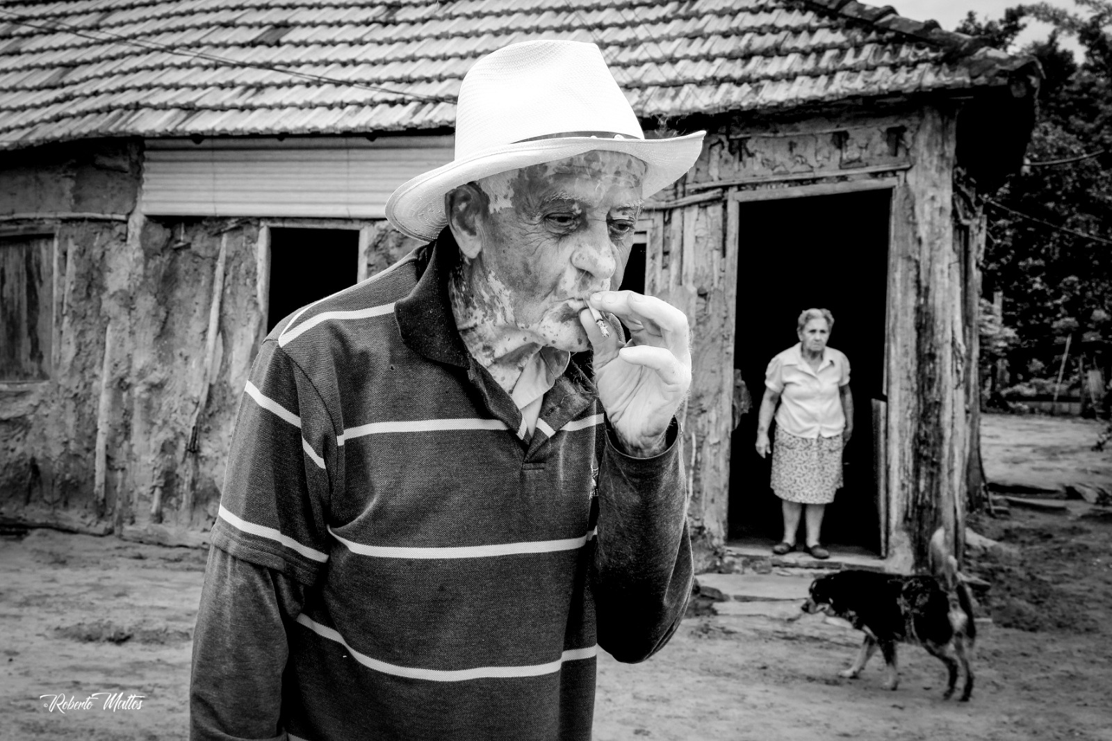 Homem de chapéu diante de uma casa rural, em preto e branco